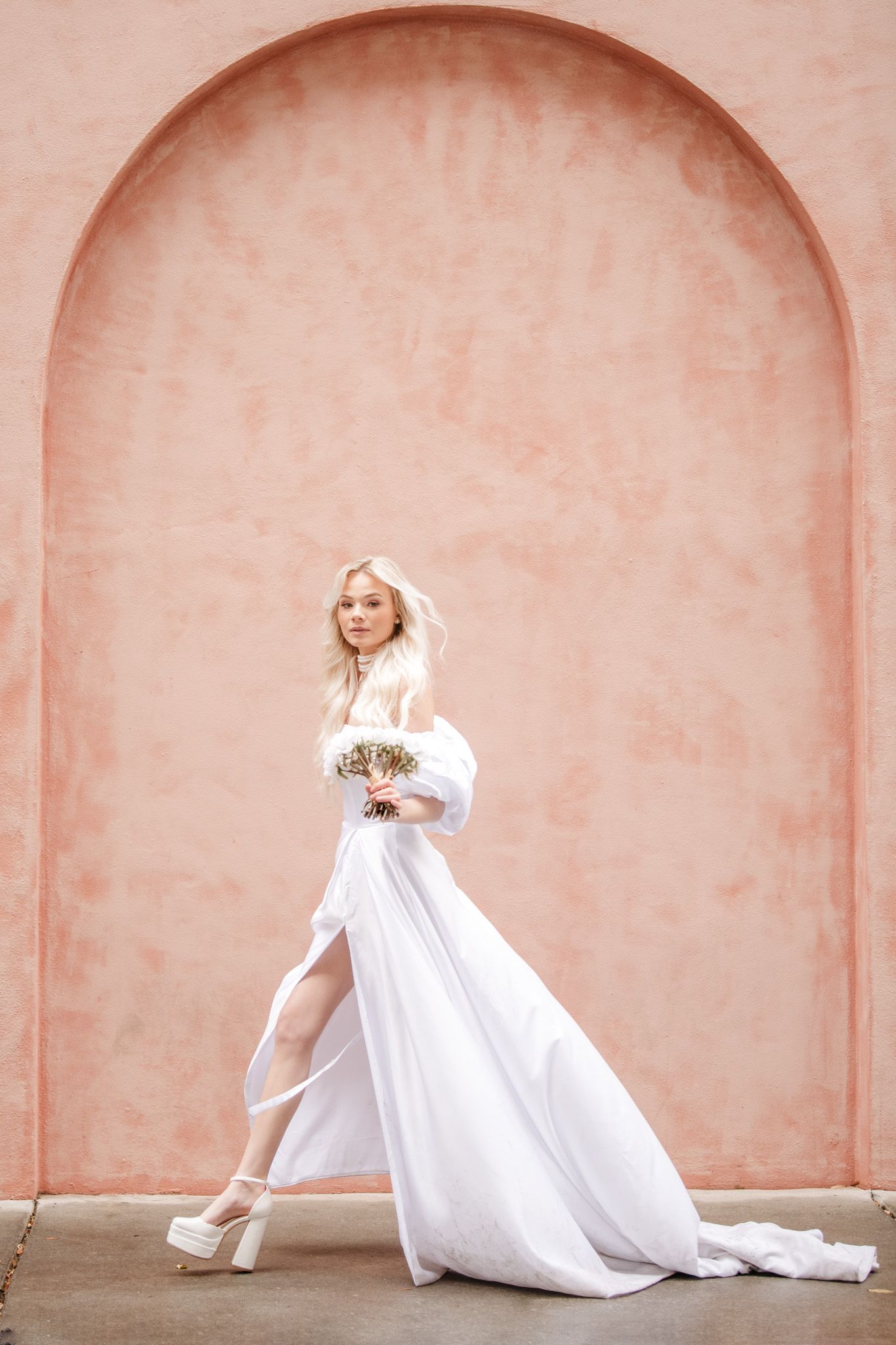 A bride in a flowing white wedding gown and heels poses elegantly against a peach-colored arched wall.