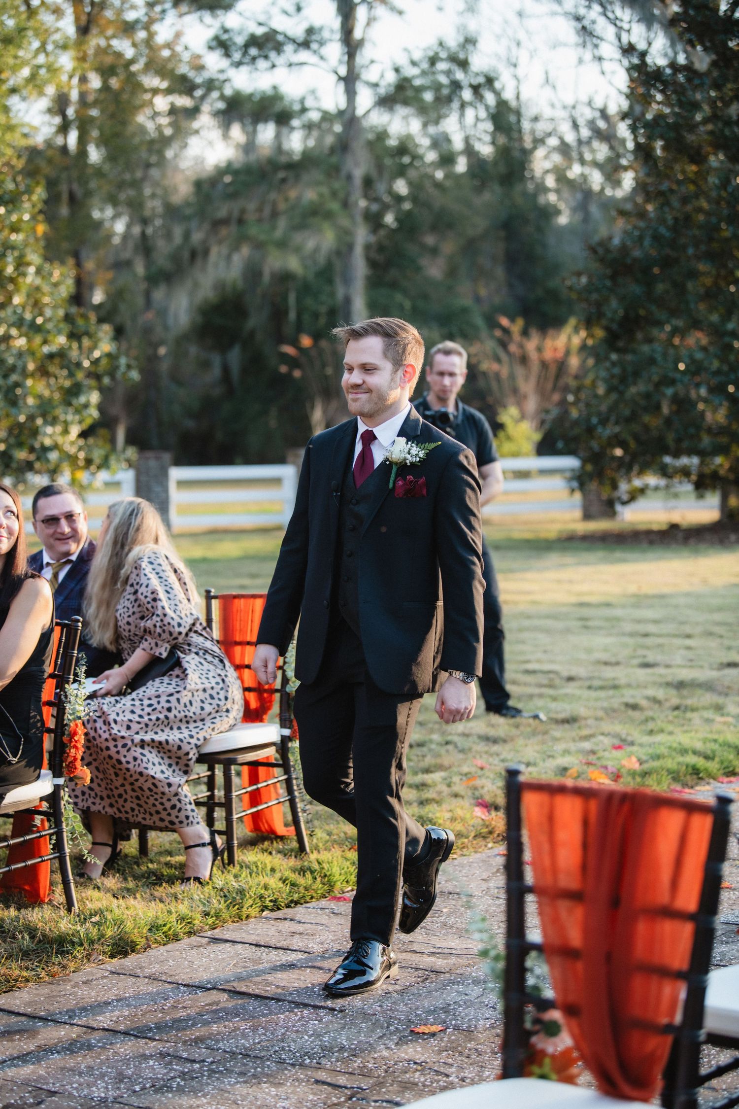 Wedding guests walk along a path with orange decor and chairs set up for an outdoor ceremony.