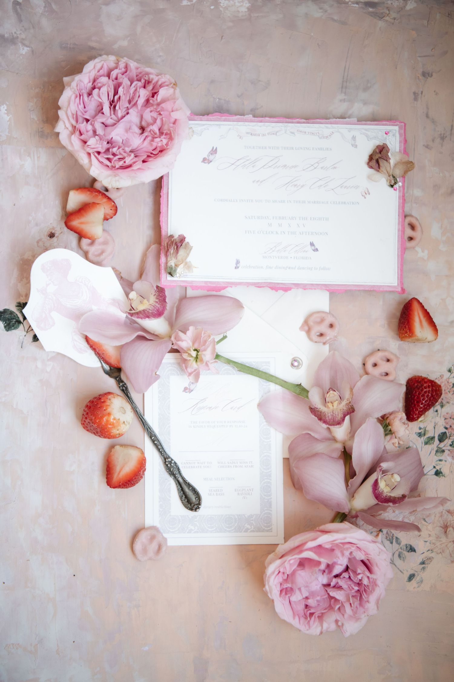 Wedding stationery flat lay with pink peonies and strawberries on white surface.