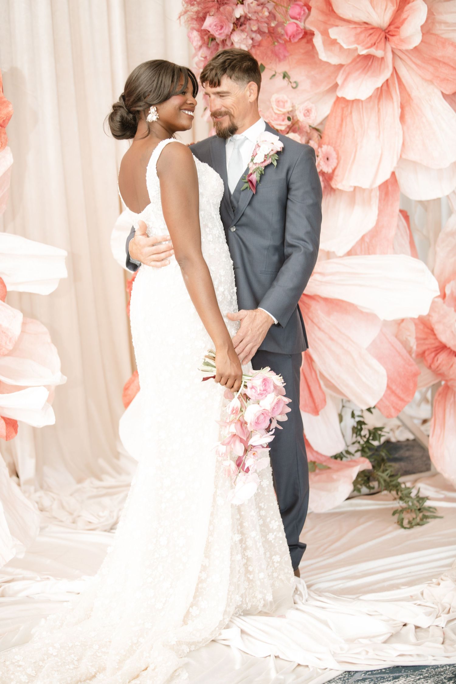 Wedding couple poses romantically against a backdrop of large coral paper flowers and white drapes.
