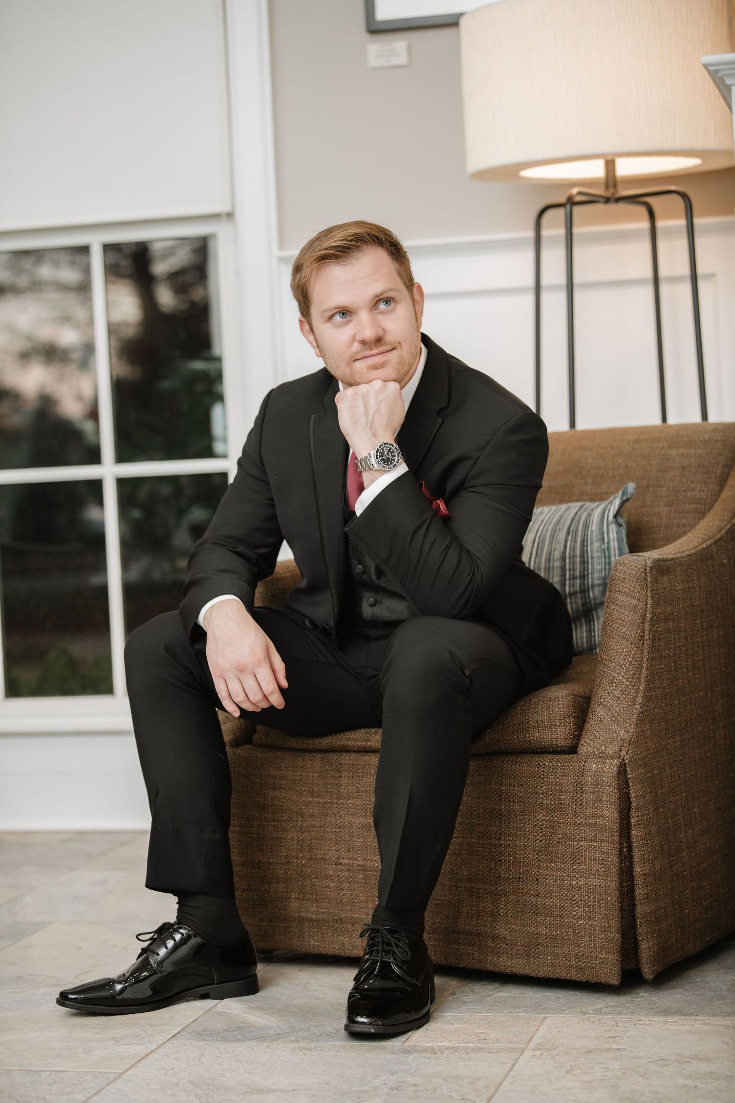 Formal portrait of person in black suit sitting thoughtfully on brown couch.