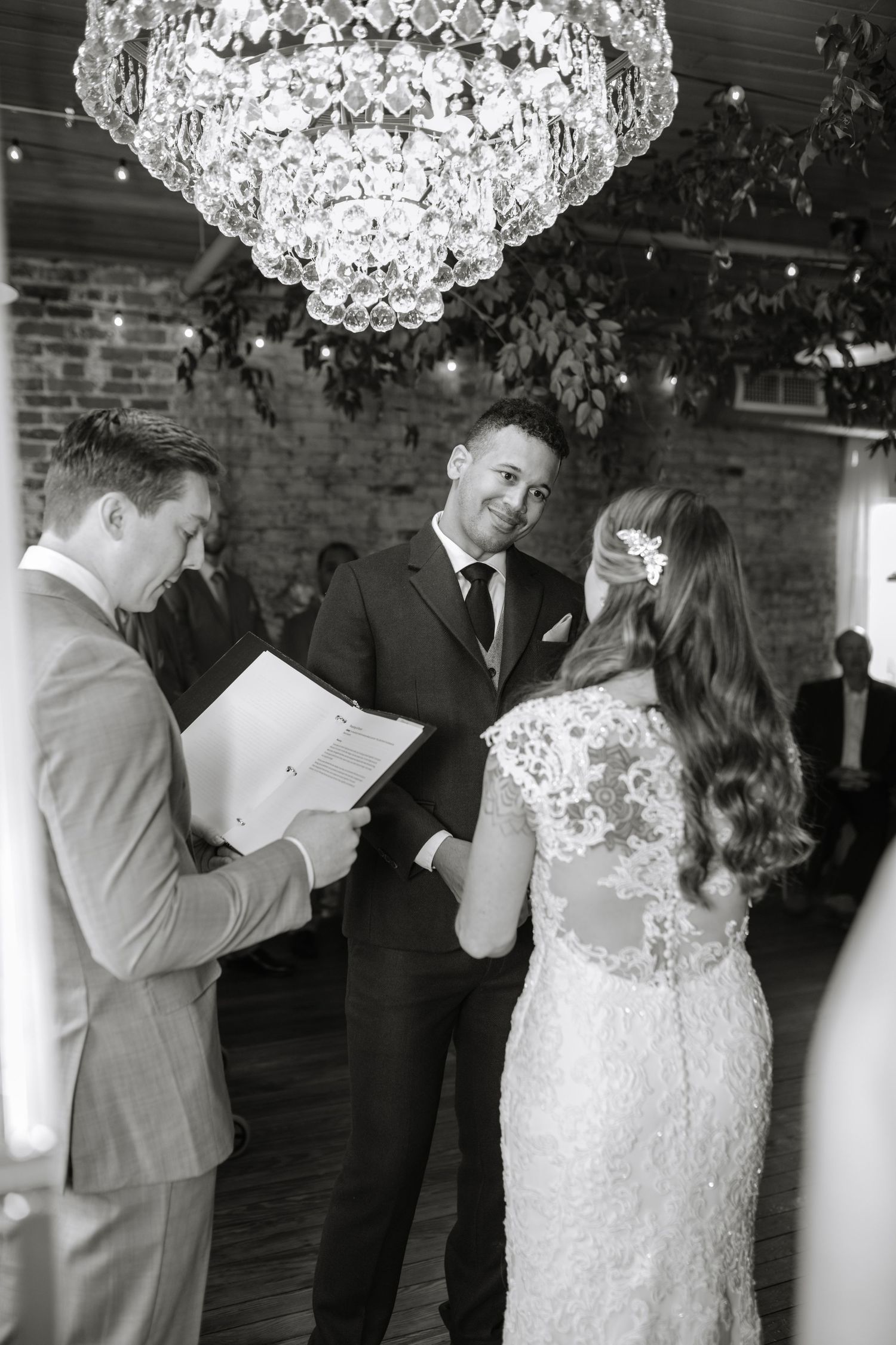 Wedding ceremony under an ornate crystal chandelier in a rustic brick-walled venue.