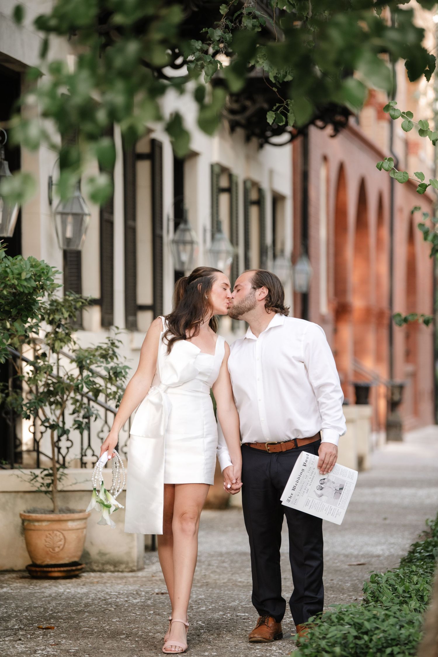 Romantic couple in white and black outfits walking and kissing in a series of photos along a street with potted plants.