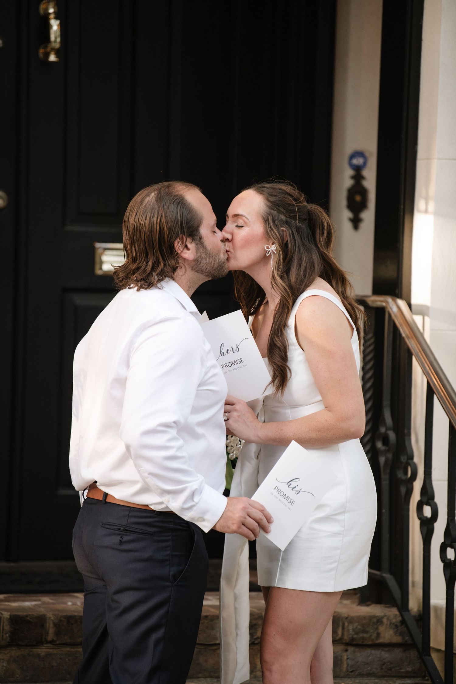 A couple in white outfits share a romantic kiss on the steps of a building.