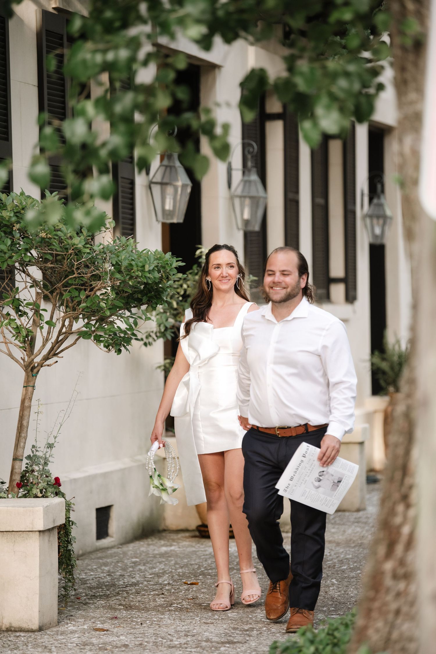 A couple in white shirts walks together along a charming cobblestone path lined with trees and lanterns.