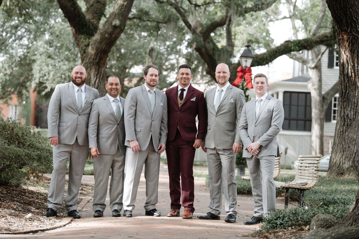 A wedding party in gray suits and burgundy attire poses together outdoors for formal group photos.