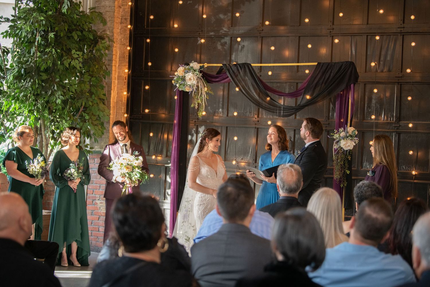 Wedding ceremony with guests watching in a dimly lit venue with twinkle lights and greenery.