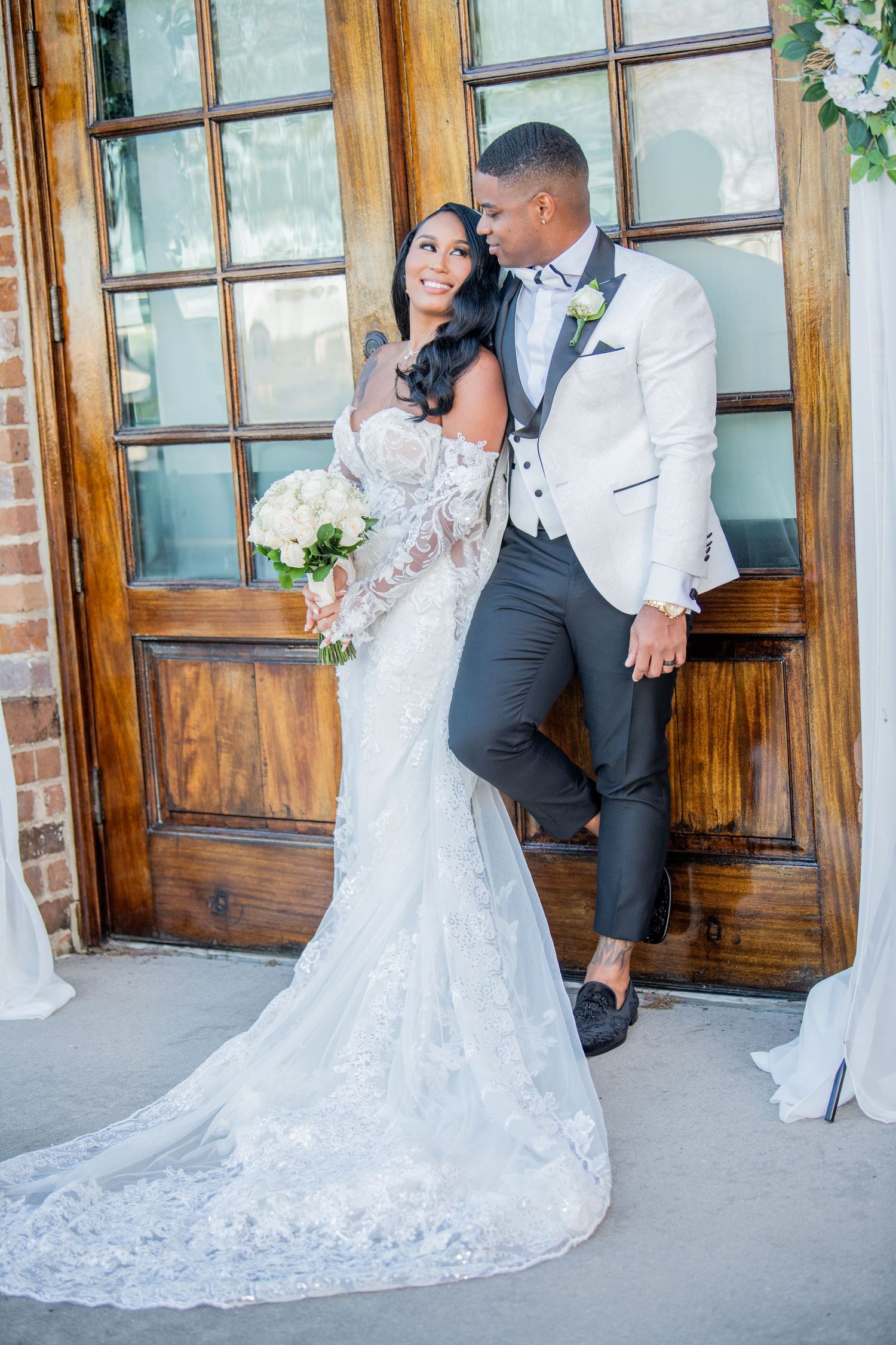 Romantic wedding portraits with couple in white and black attire by wooden doors and brick wall.