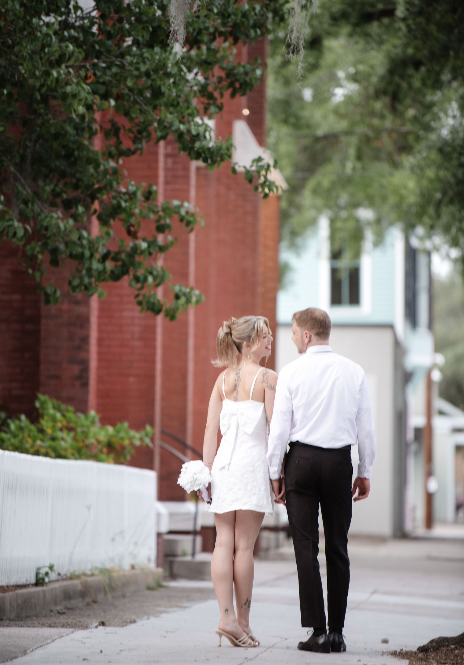A couple in formal attire walking down a sidewalk next to a red brick building.