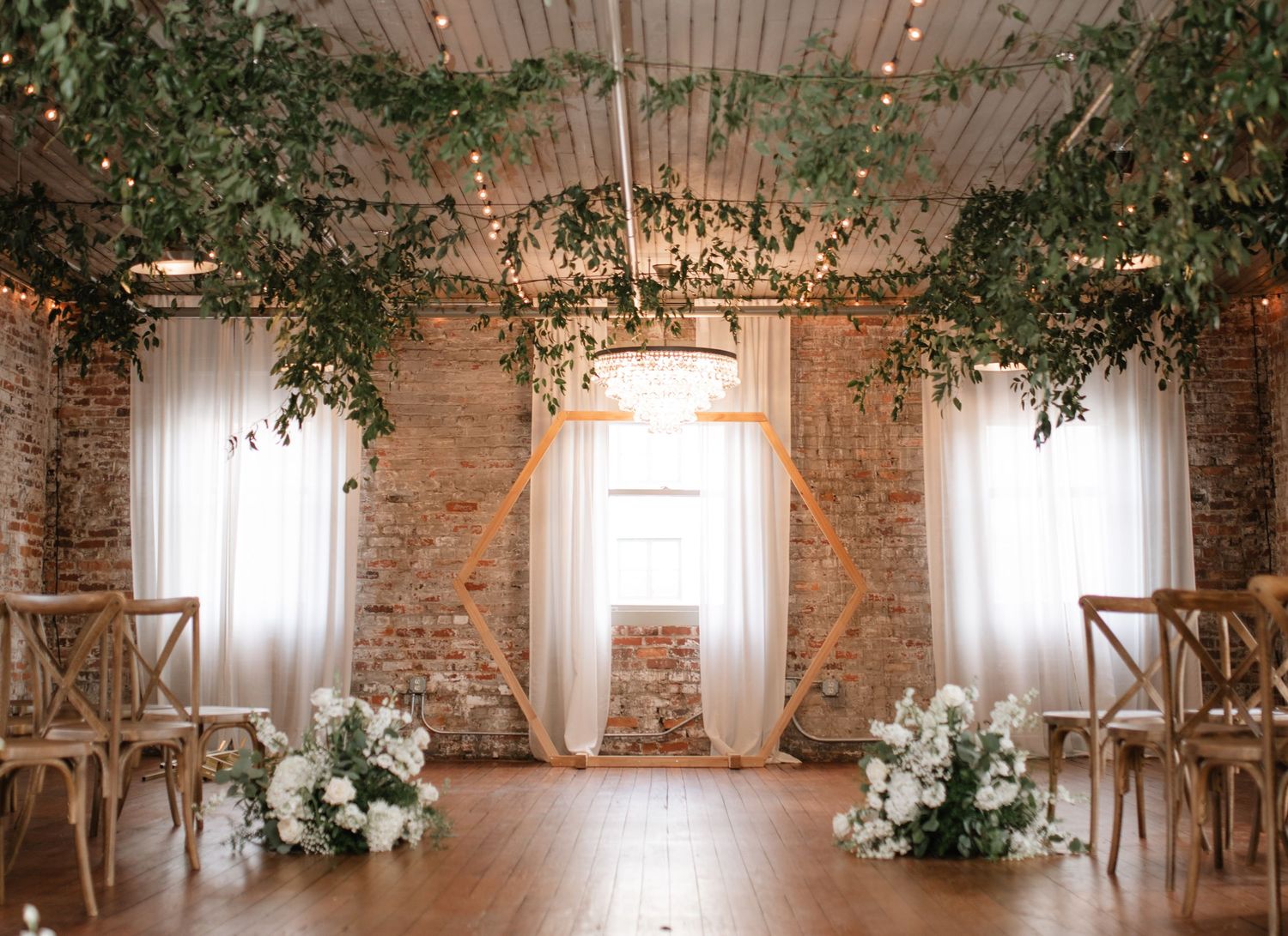 Elegant wedding venue interior with exposed brick walls, hanging greenery, and natural light streaming through windows.