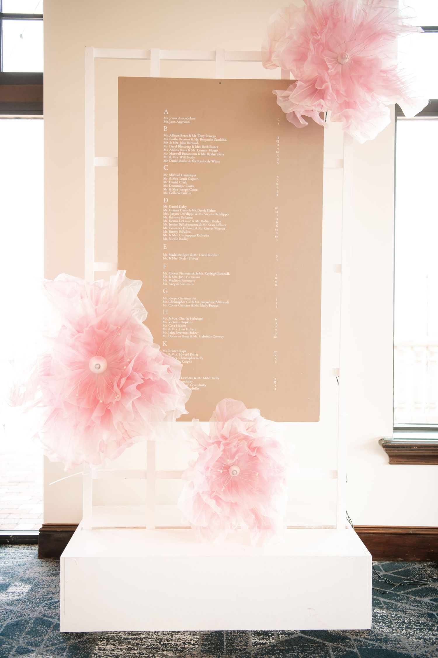 Modern wedding seating chart display with pink tissue paper pom poms on white backdrop.