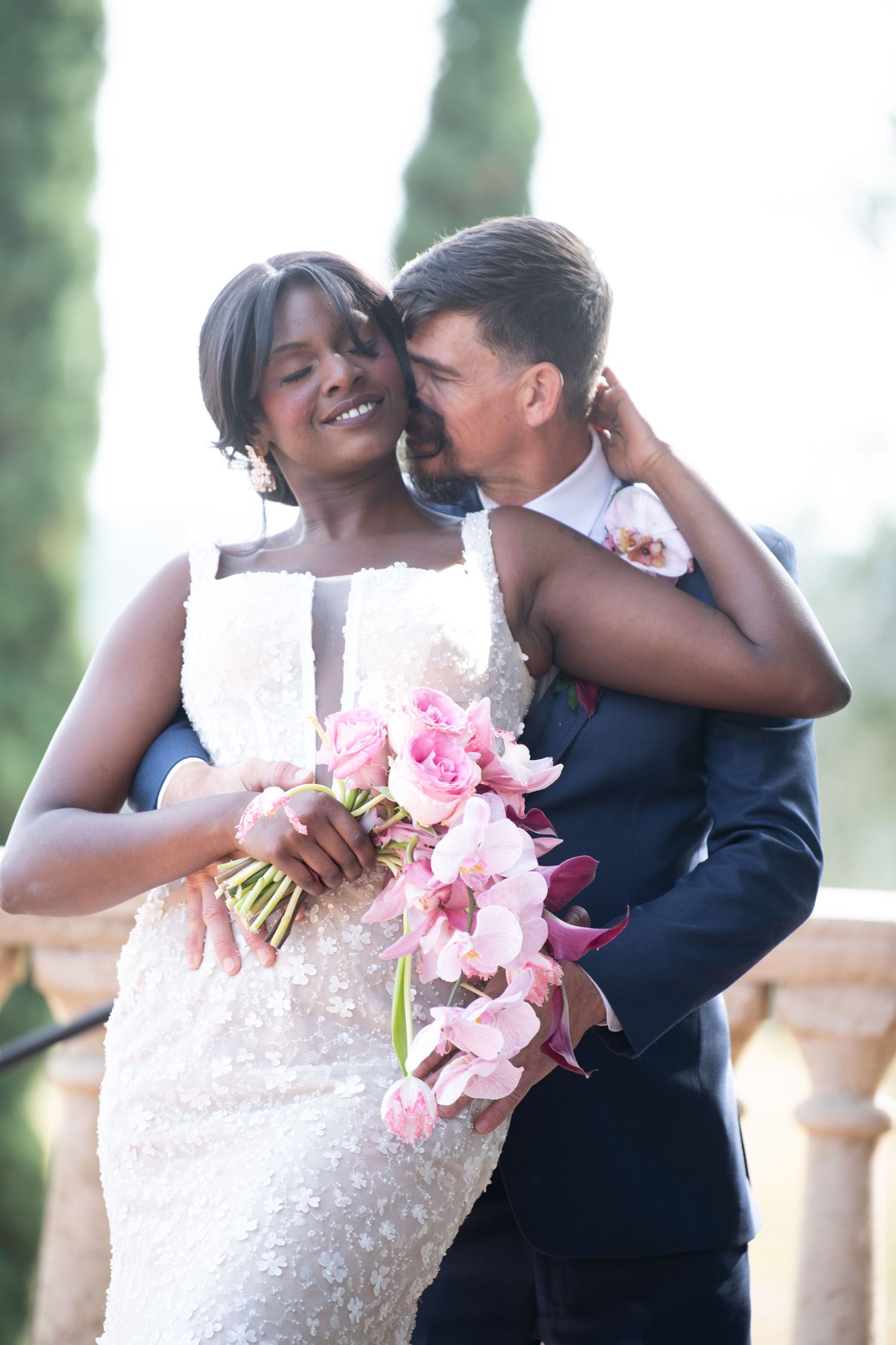 Newlyweds share intimate moments while holding pink orchids during outdoor photo session.
