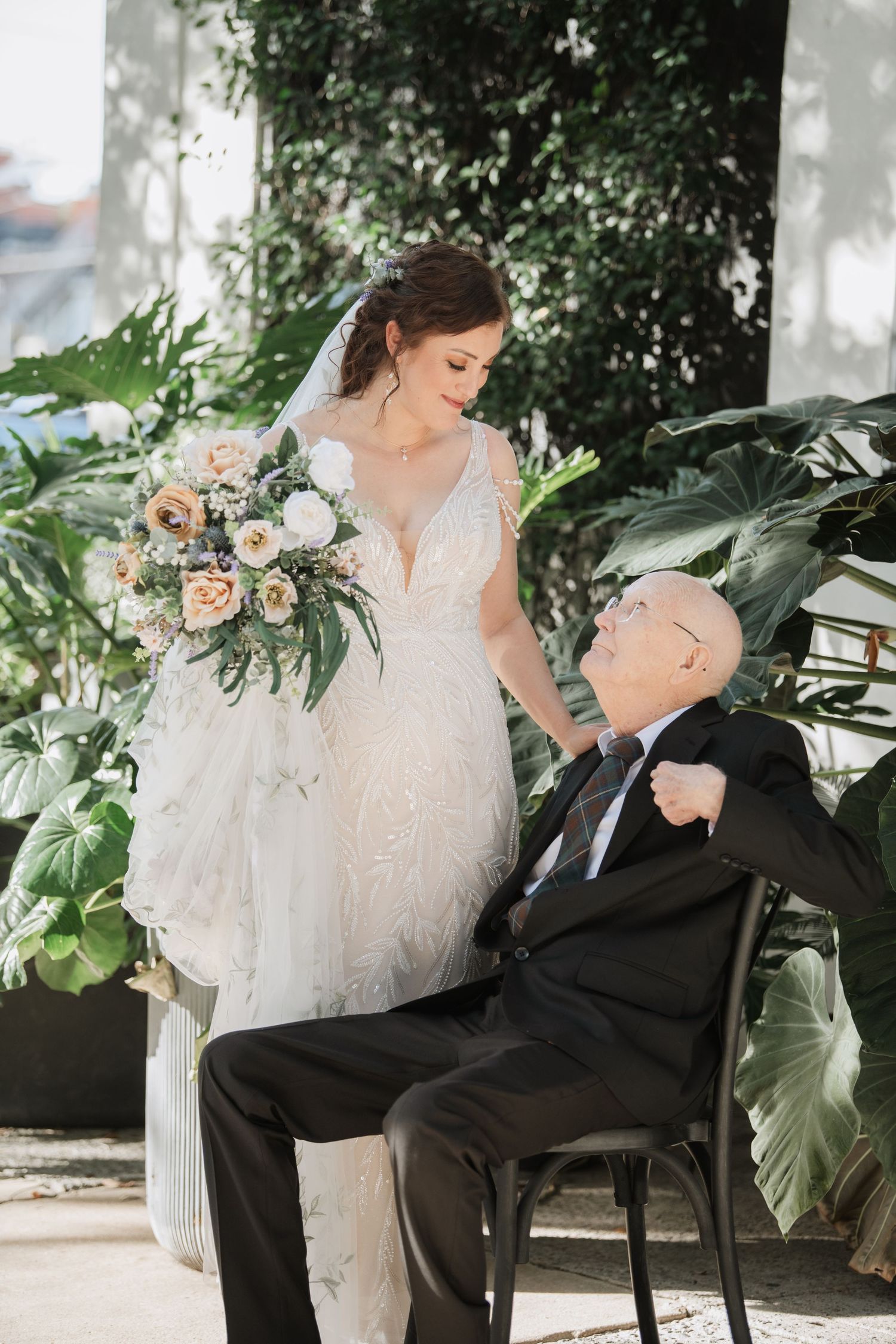 Tender moment between bride in lace gown and elderly guest in wheelchair among lush greenery.