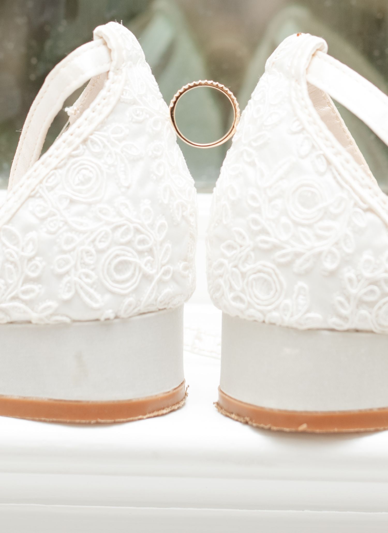 Close-up of white lace wedding flats with gold wedding band balanced between them.