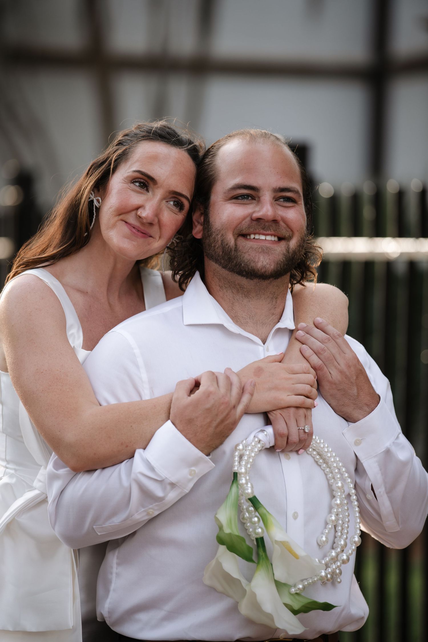 A couple embraces outdoors in wedding attire, with the person in white holding a small decorative handbag.