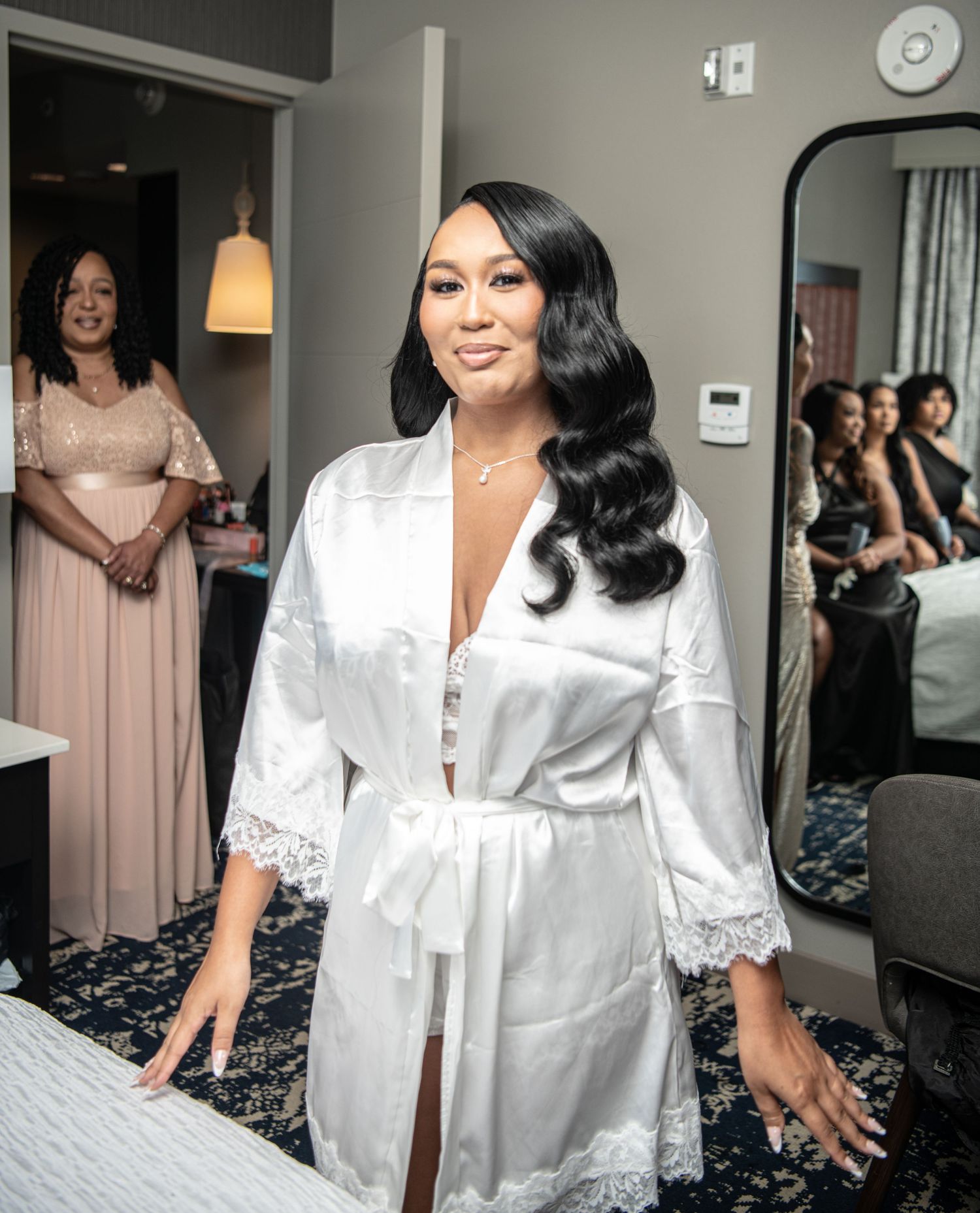 Bride in white silk robe with lace details preparing in hotel room on wedding morning.