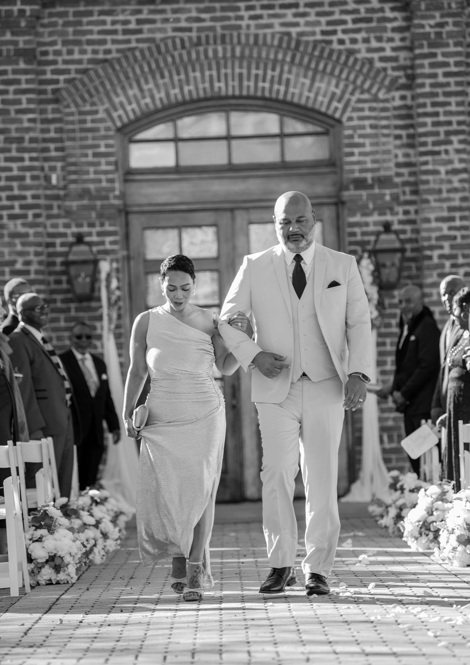 Black and white photo of wedding party members walking down brick-lined aisle with floral decor.