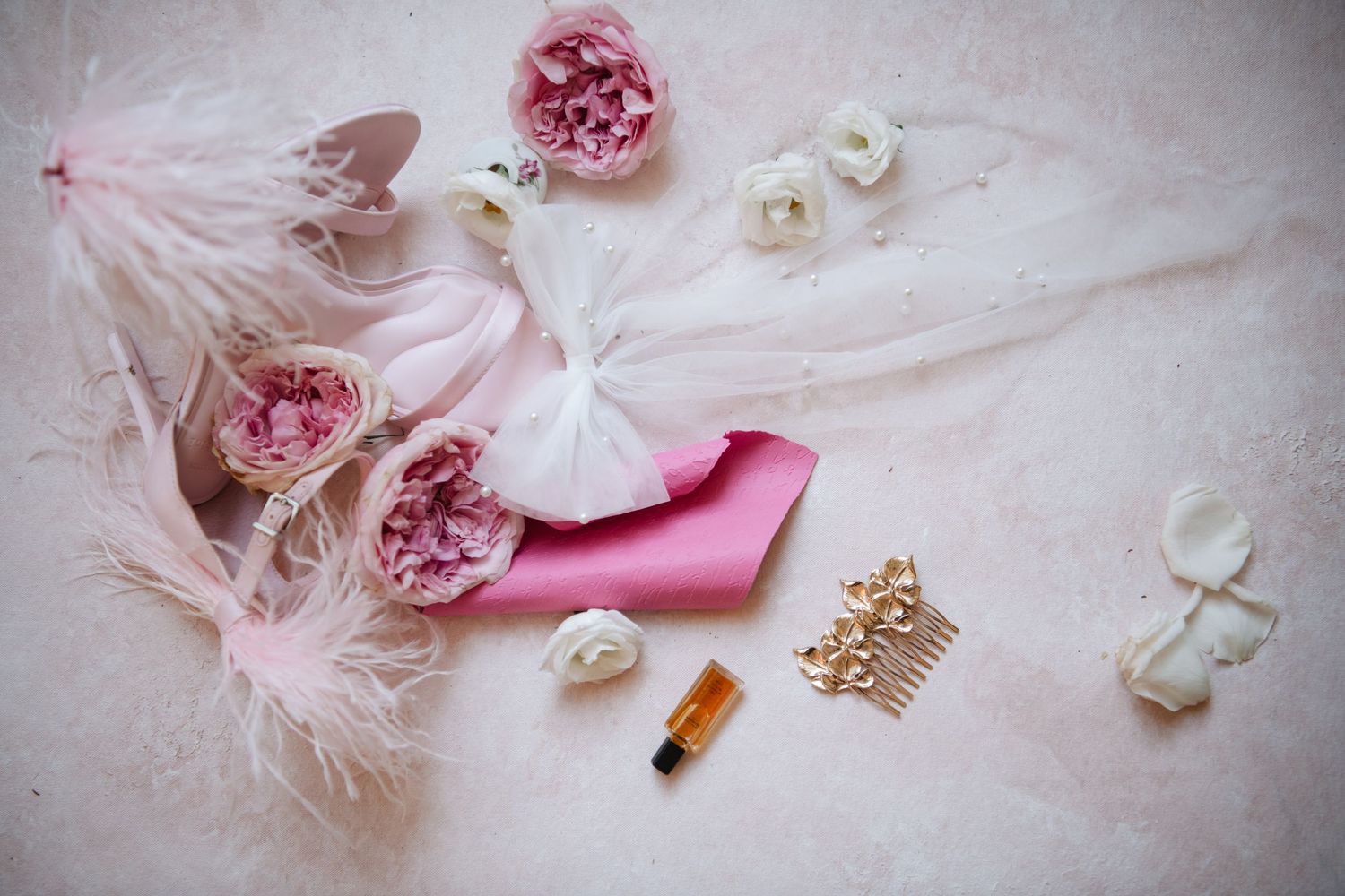 Wedding accessories including pink flowers, feathers, and gold crown on light pink surface.