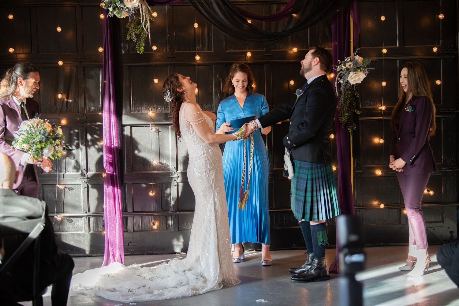 Wedding ceremony in industrial venue with string lights and purple fabric draping.