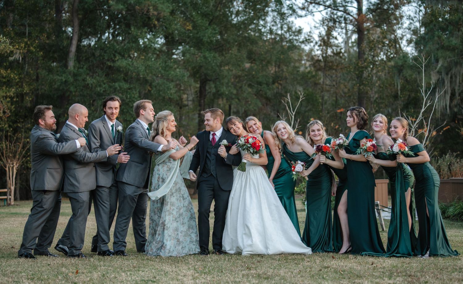 Wedding party poses together outdoors in formal attire with bridesmaids in emerald green dresses.