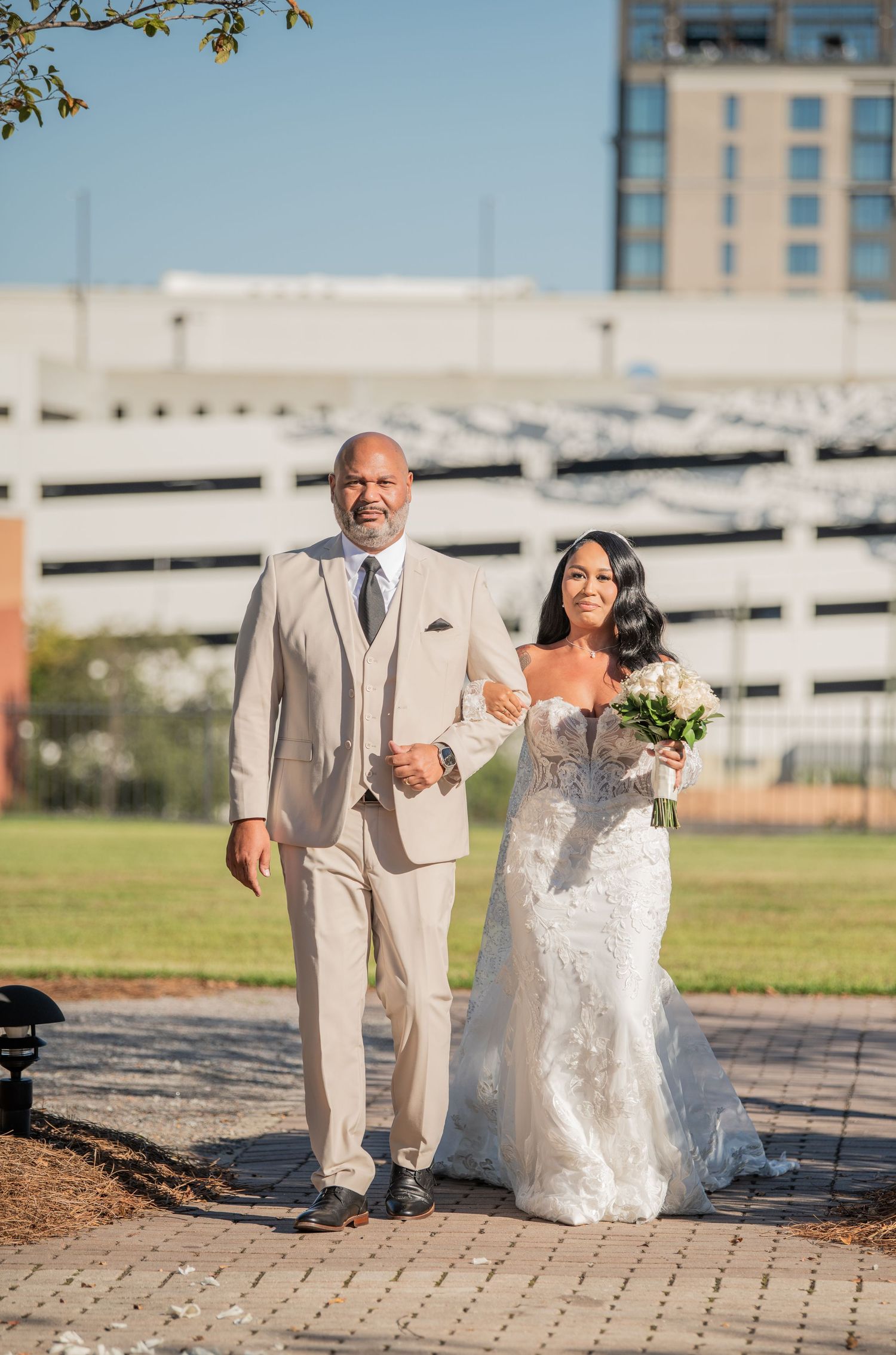 A wedding couple walks along a paved pathway with an urban backdrop.