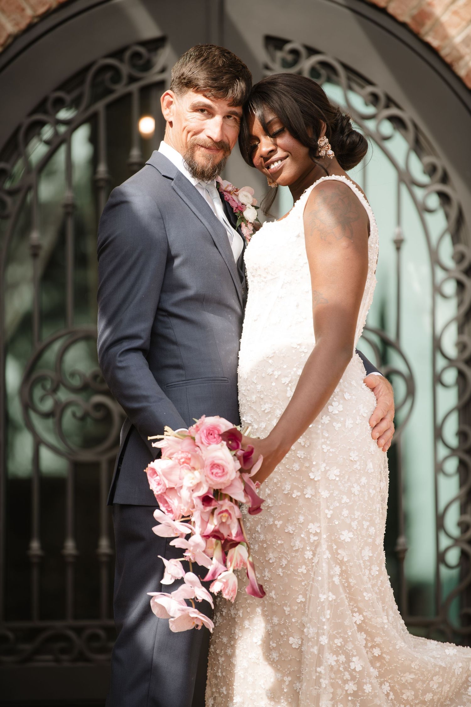 A couple in wedding attire embraces near an ornate stone archway with a pink floral bouquet.