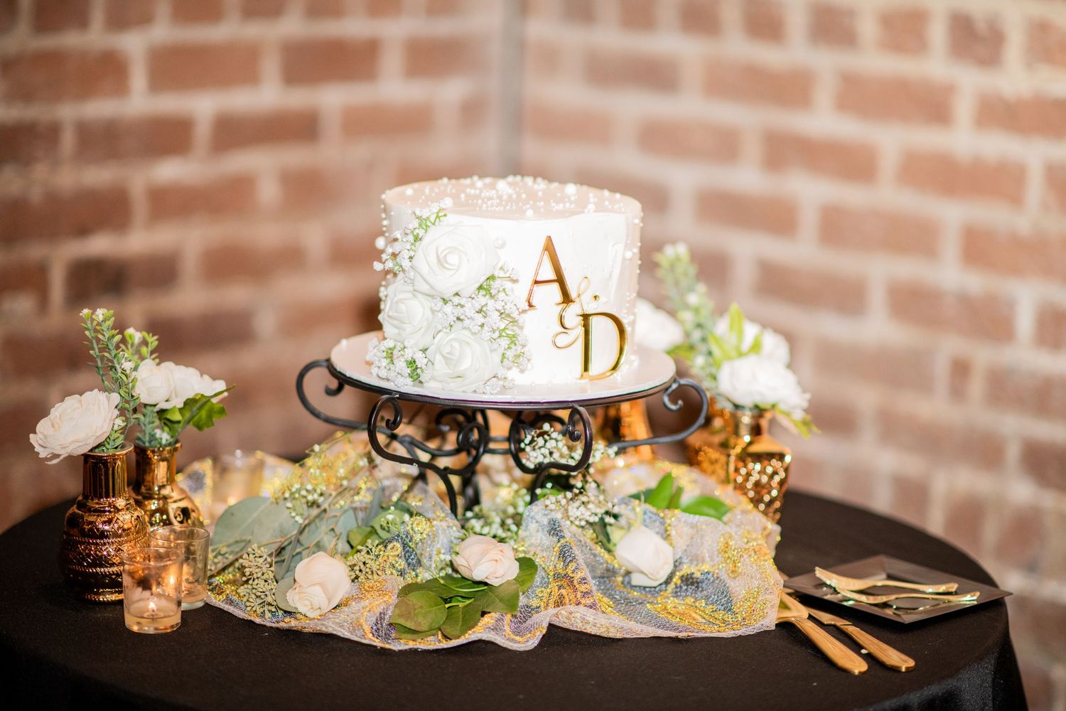 Elegant white wedding cake with monogram initials decorated with delicate flowers on a black iron stand.