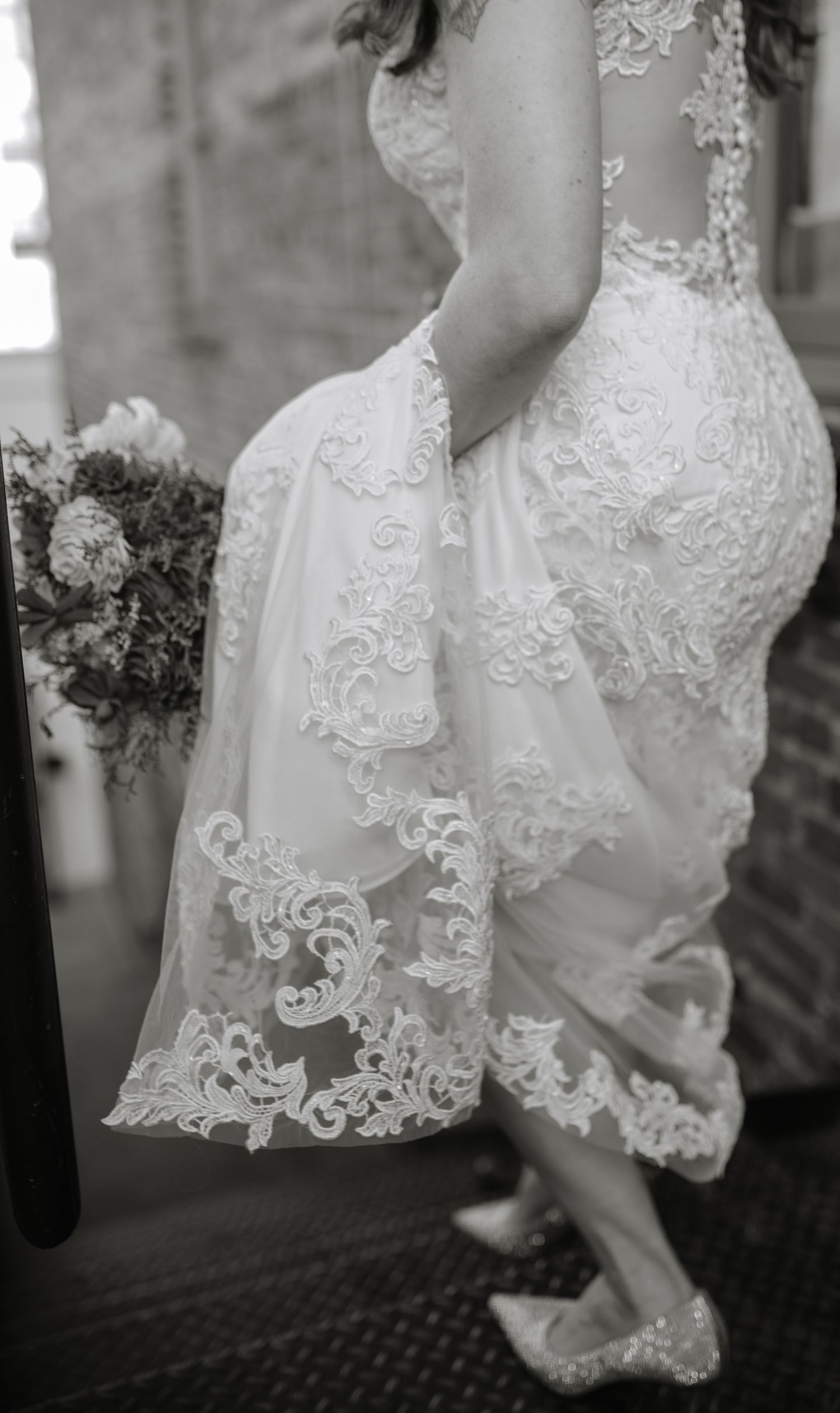 Black and white photo showing intricate lace details of a wedding dress and bouquet.