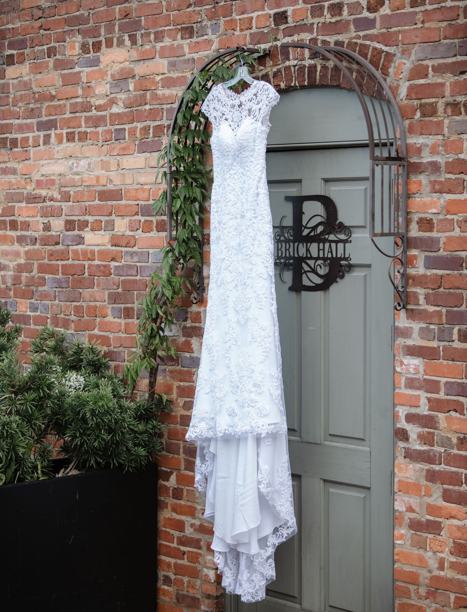 White lace wedding dress hangs against rustic brick wall with grey door.
