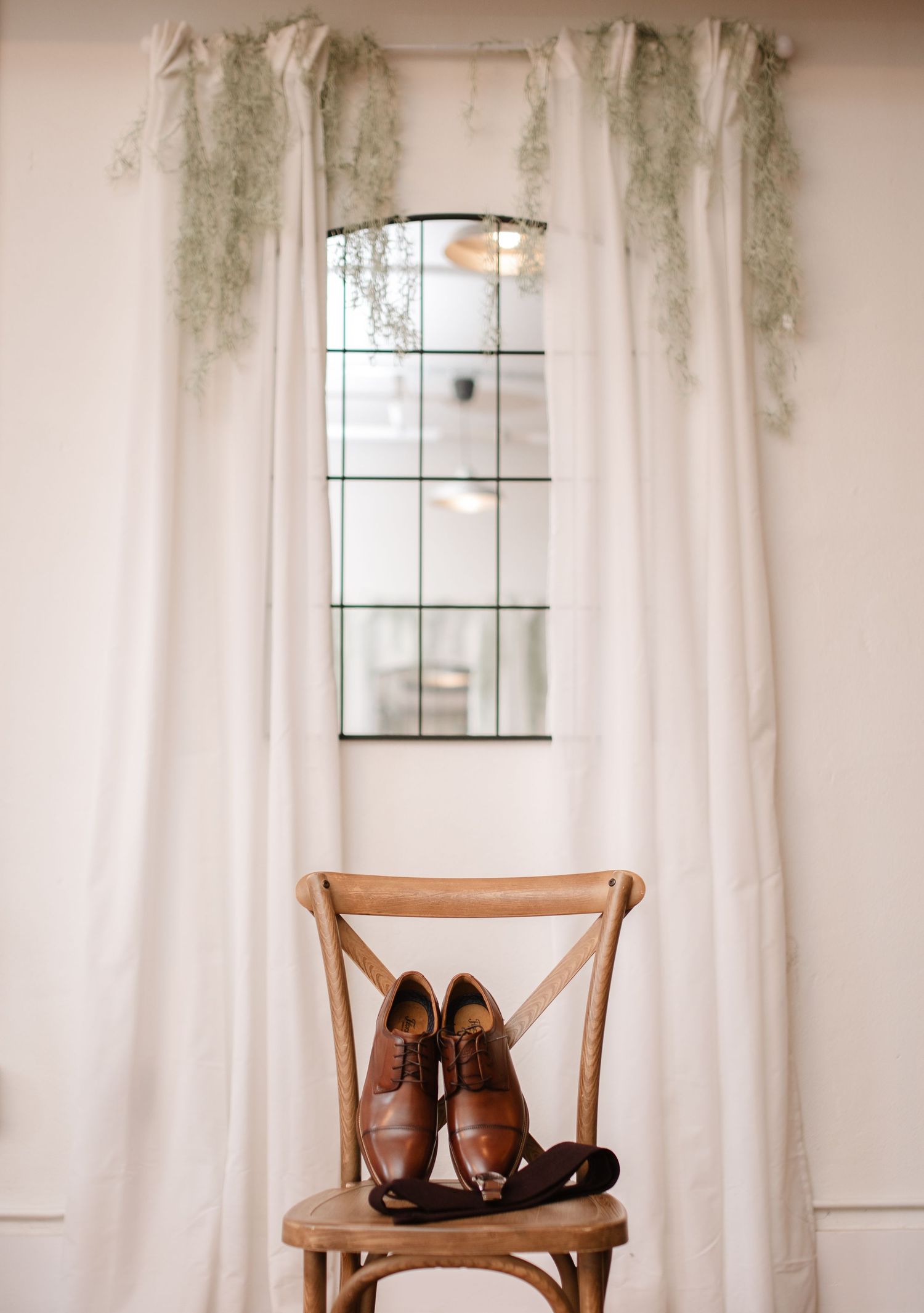 Brown leather dress shoes displayed on wooden chair against white curtained window.
