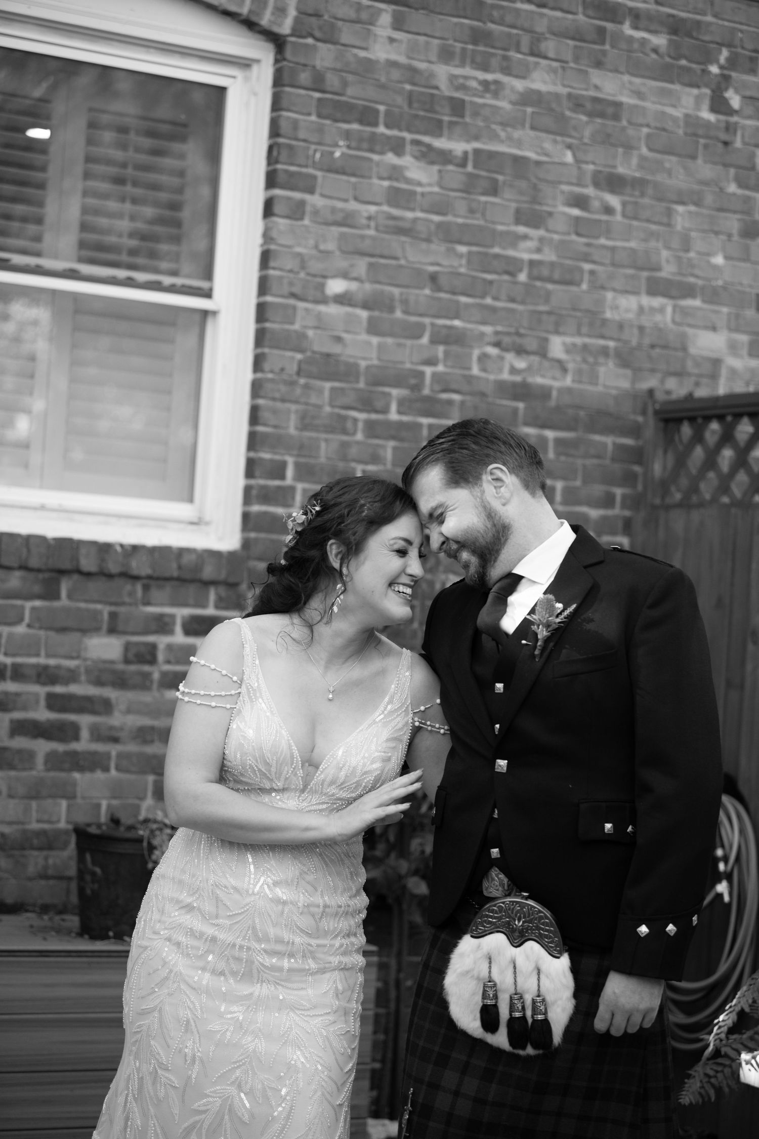 Intimate black and white portrait of newlyweds against brick wall with groom wearing traditional Scottish kilt.