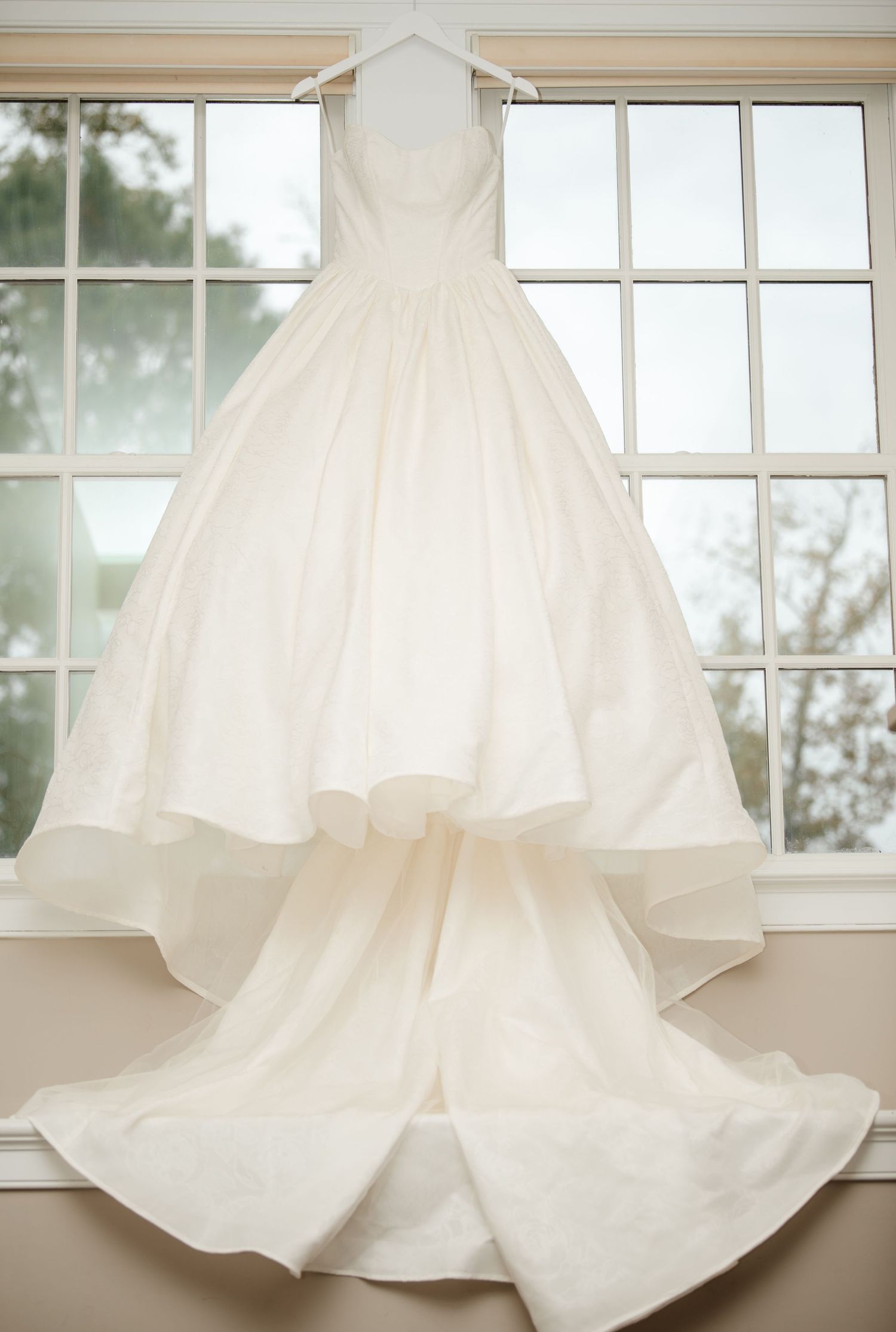 Elegant white wedding gown with full skirt displayed against window backdrop.