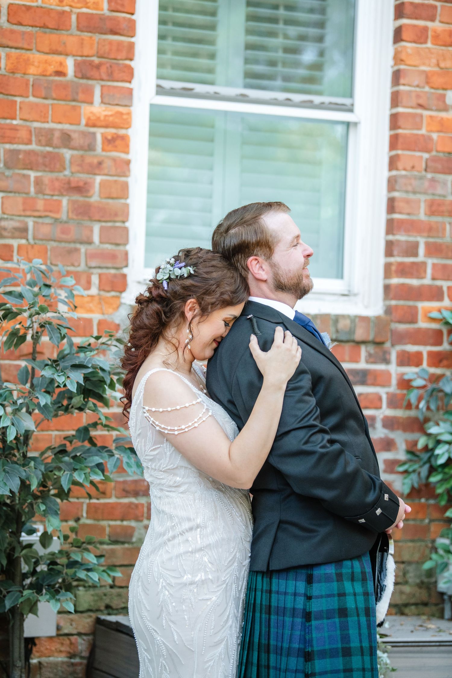 Tender embrace between couple in wedding attire against rustic brick backdrop with window.