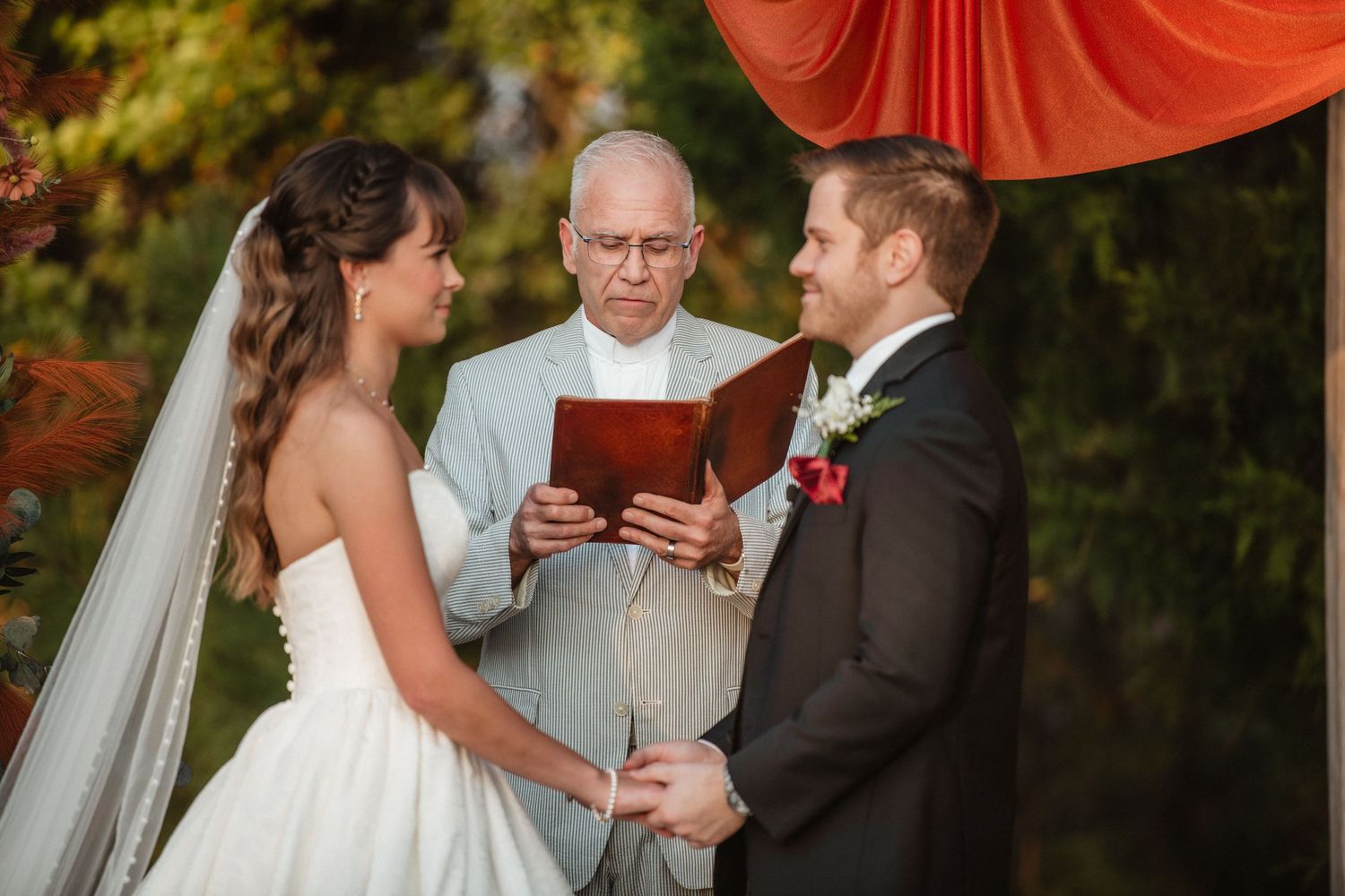 A wedding ceremony takes place under an orange draped arch decorated with floral arrangements.
