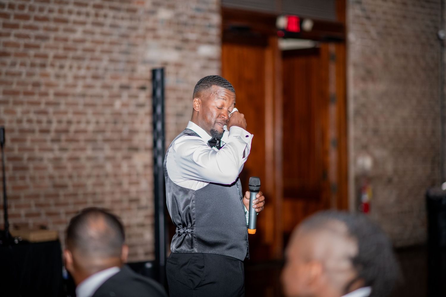 A speaker in a grey vest holds a microphone during a reception at a brick-walled venue.