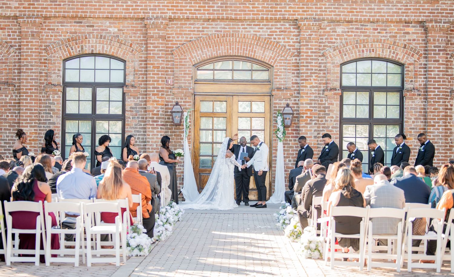 Wide panoramic shot of wedding ceremony setup with white chairs and brick backdrop.