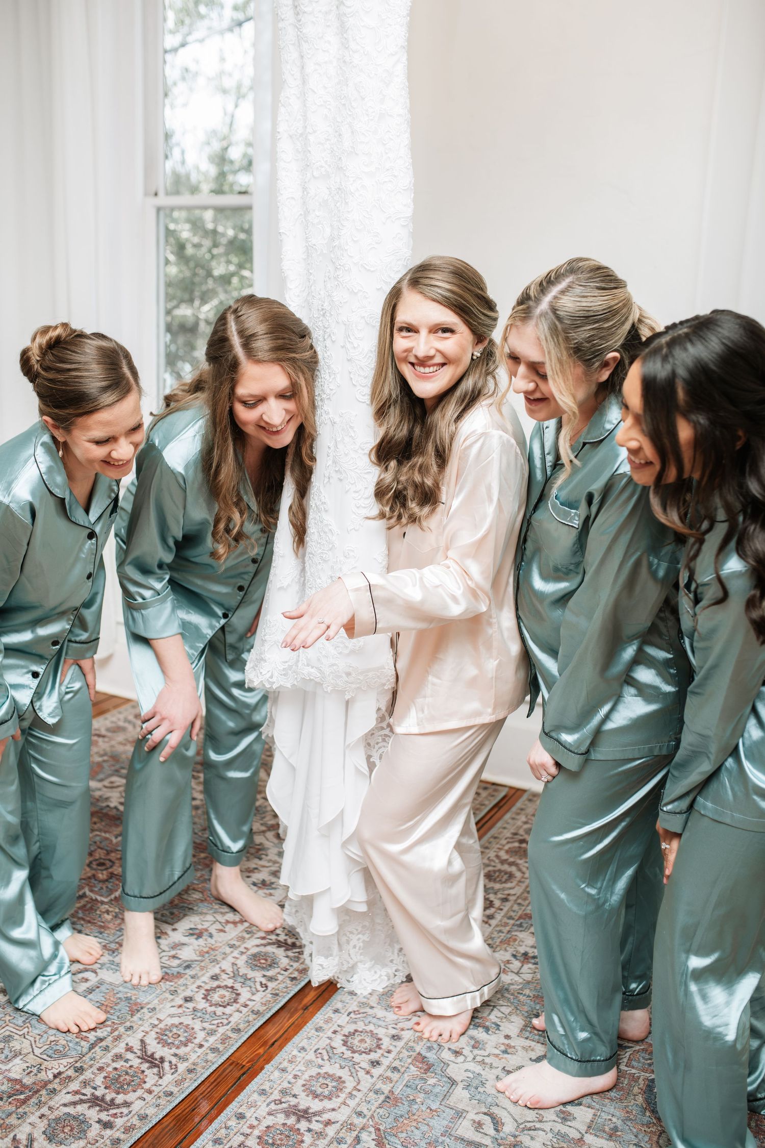 Bridesmaids in matching sage robes laugh together during wedding preparations.