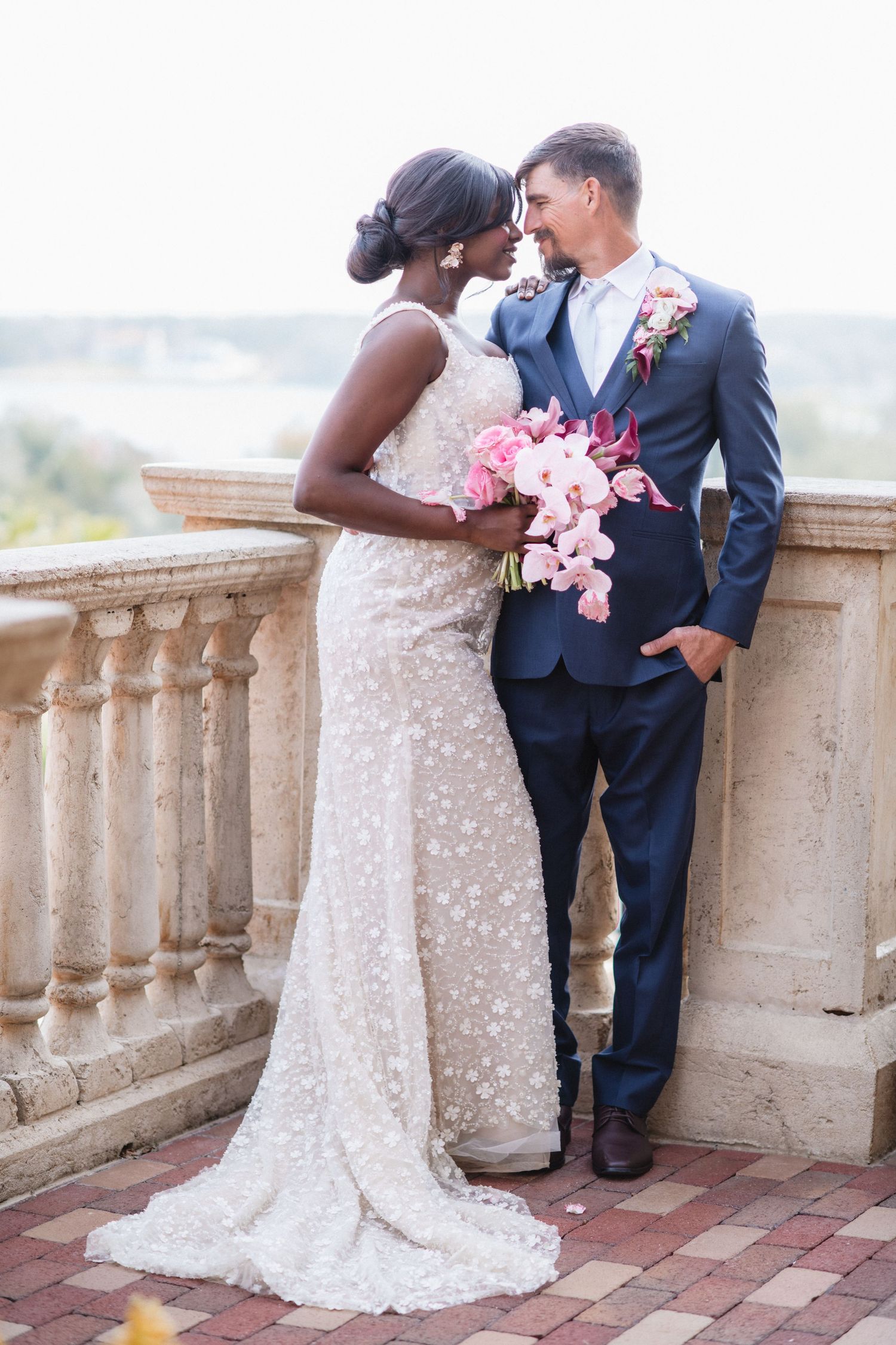 Romantic couple in wedding attire standing by stone balustrade.