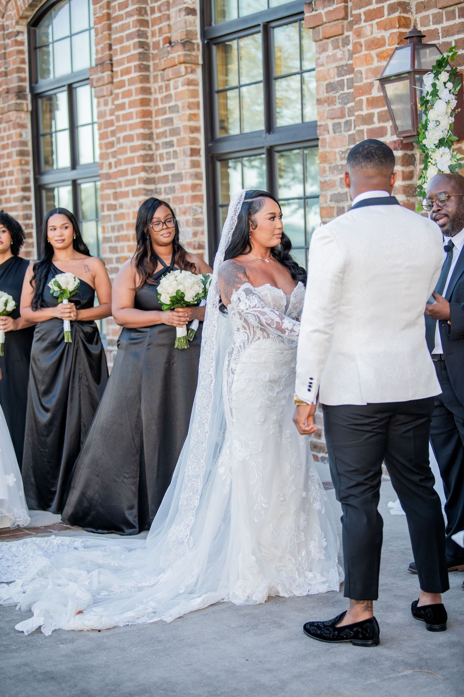 Wedding party in formal attire poses against brick wall with bridesmaids in dark dresses.