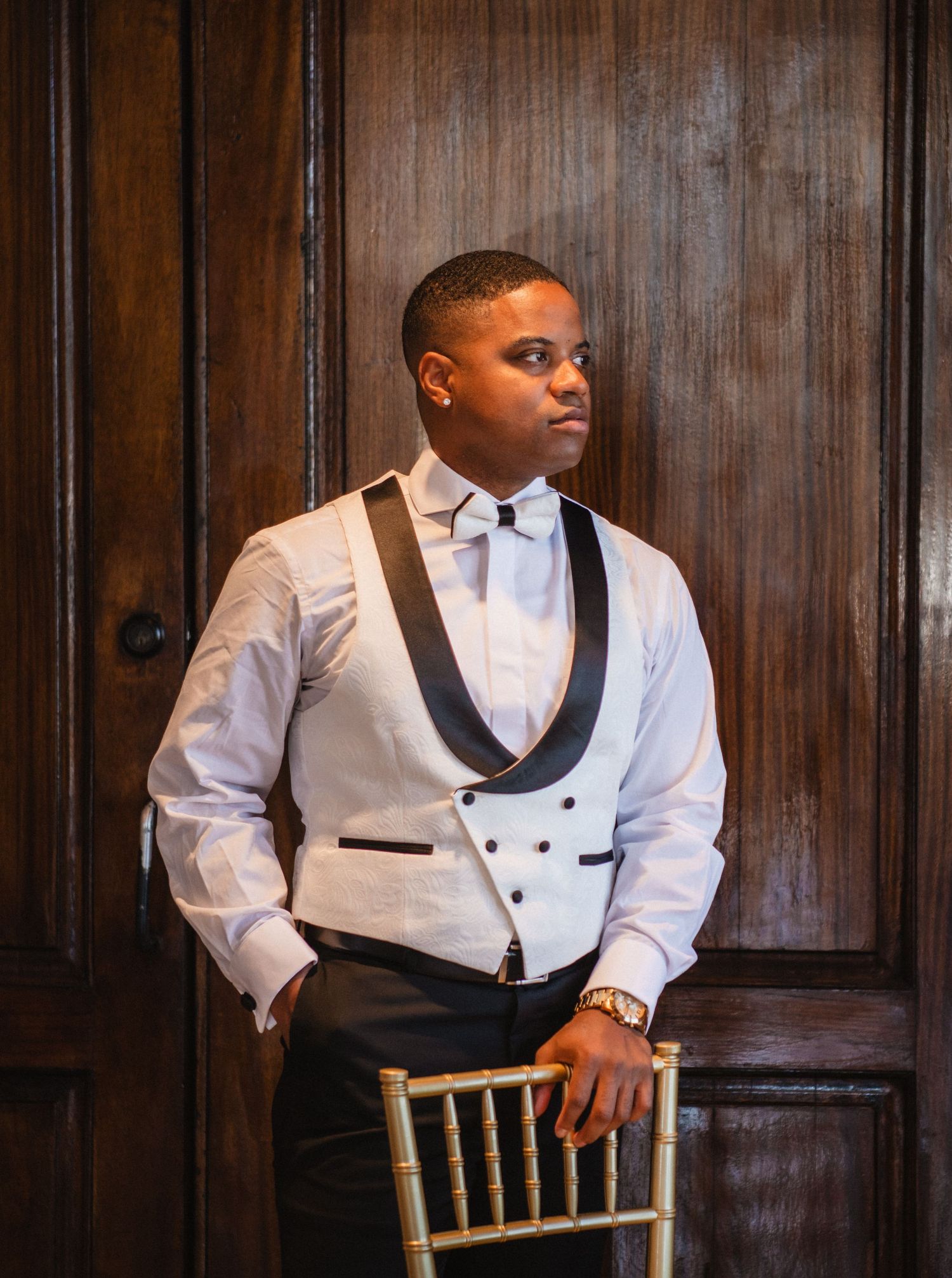 Groomsman in white dinner jacket sits on wooden chair against dark wood paneling.