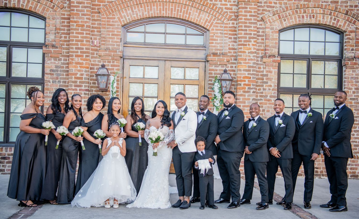 Panoramic shot of wedding party in formal attire posing against brick building exterior.