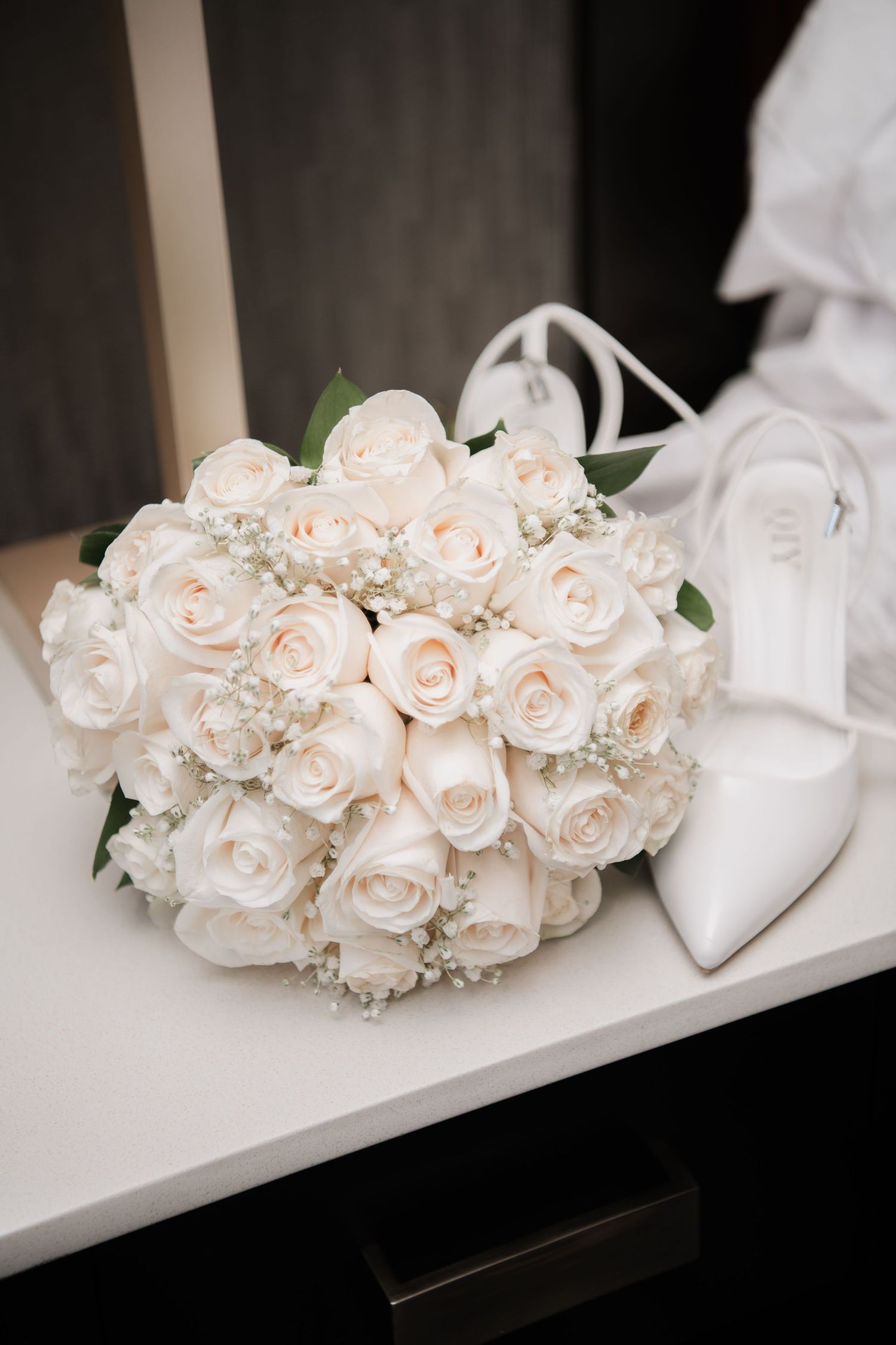 Delicate blush pink and white roses arranged in a bridal bouquet next to white heels.