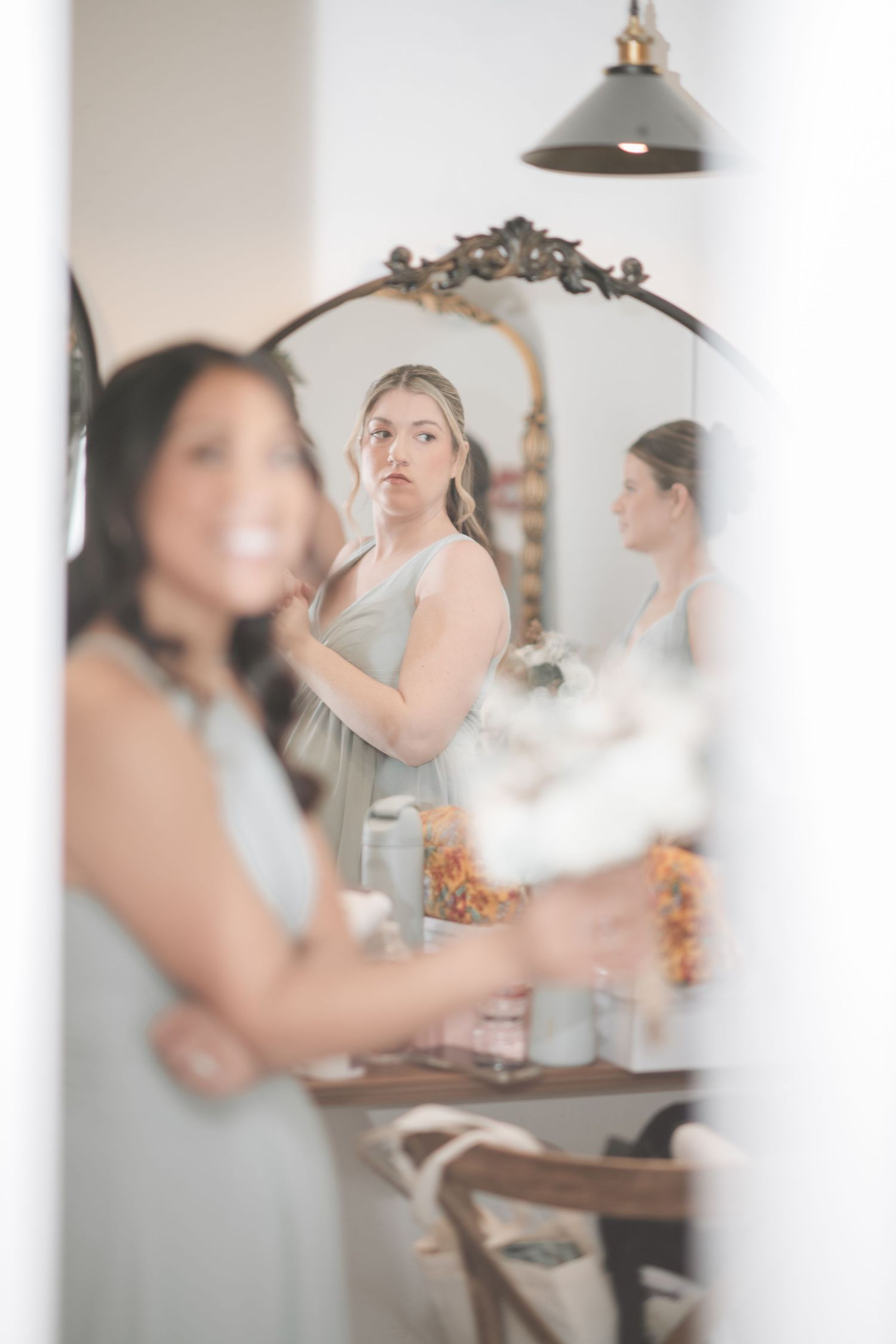 A dreamy reflection in an ornate mirror shows people getting ready in a softly lit room with a pendant lamp.
