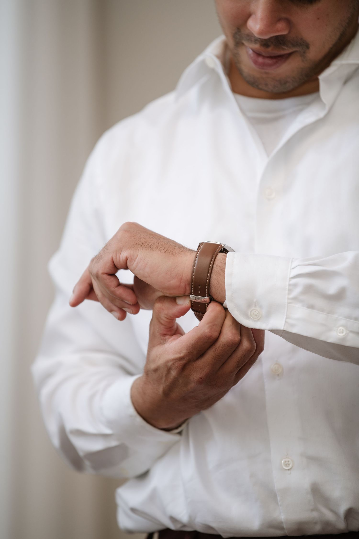 Close-up of hands adjusting brown leather watch strap on white dress shirt sleeve.