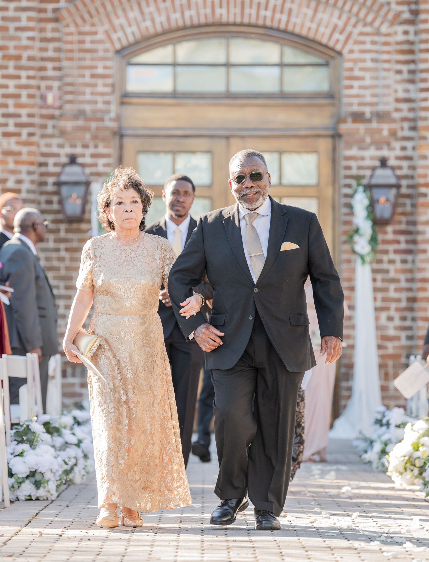 Wedding guests in formal attire walking down decorated aisle at brick venue.