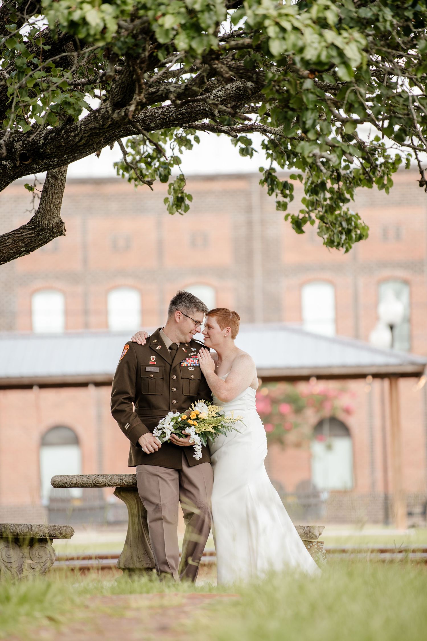 Wedding couple poses under a shady tree near a brick building.