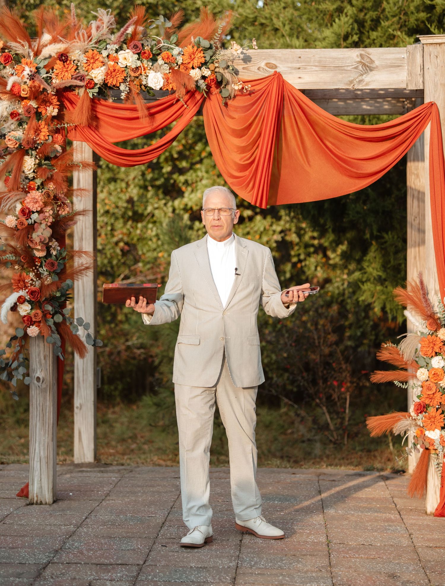 A person in a light gray suit stands under an orange draped arch decorated with flowers at an outdoor event.