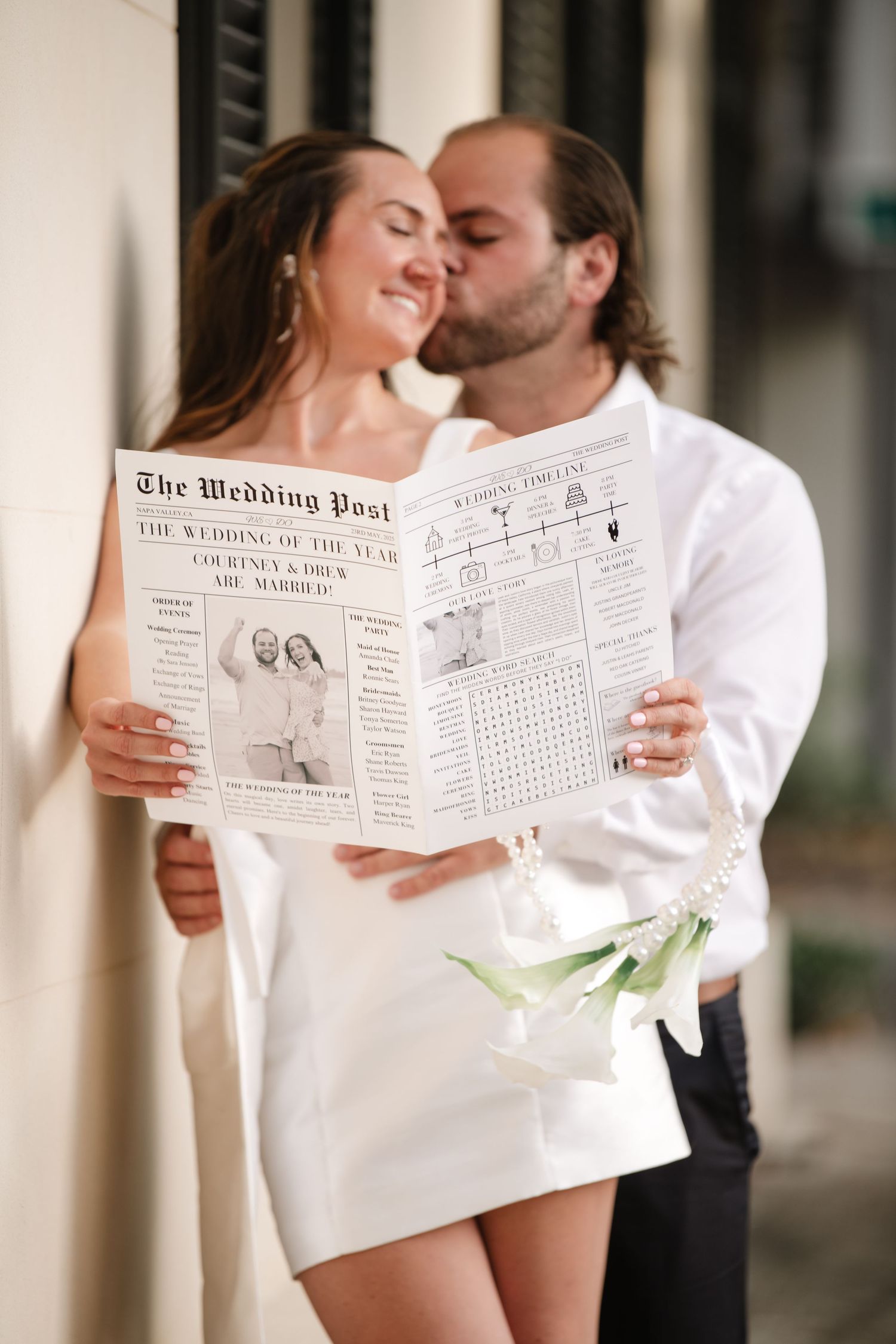 A couple shares an intimate moment while holding a wedding-themed newspaper in matching white outfits.
