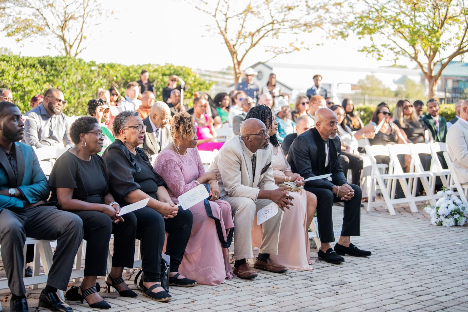 Wedding guests seated in white chairs during outdoor ceremony with trees in background.