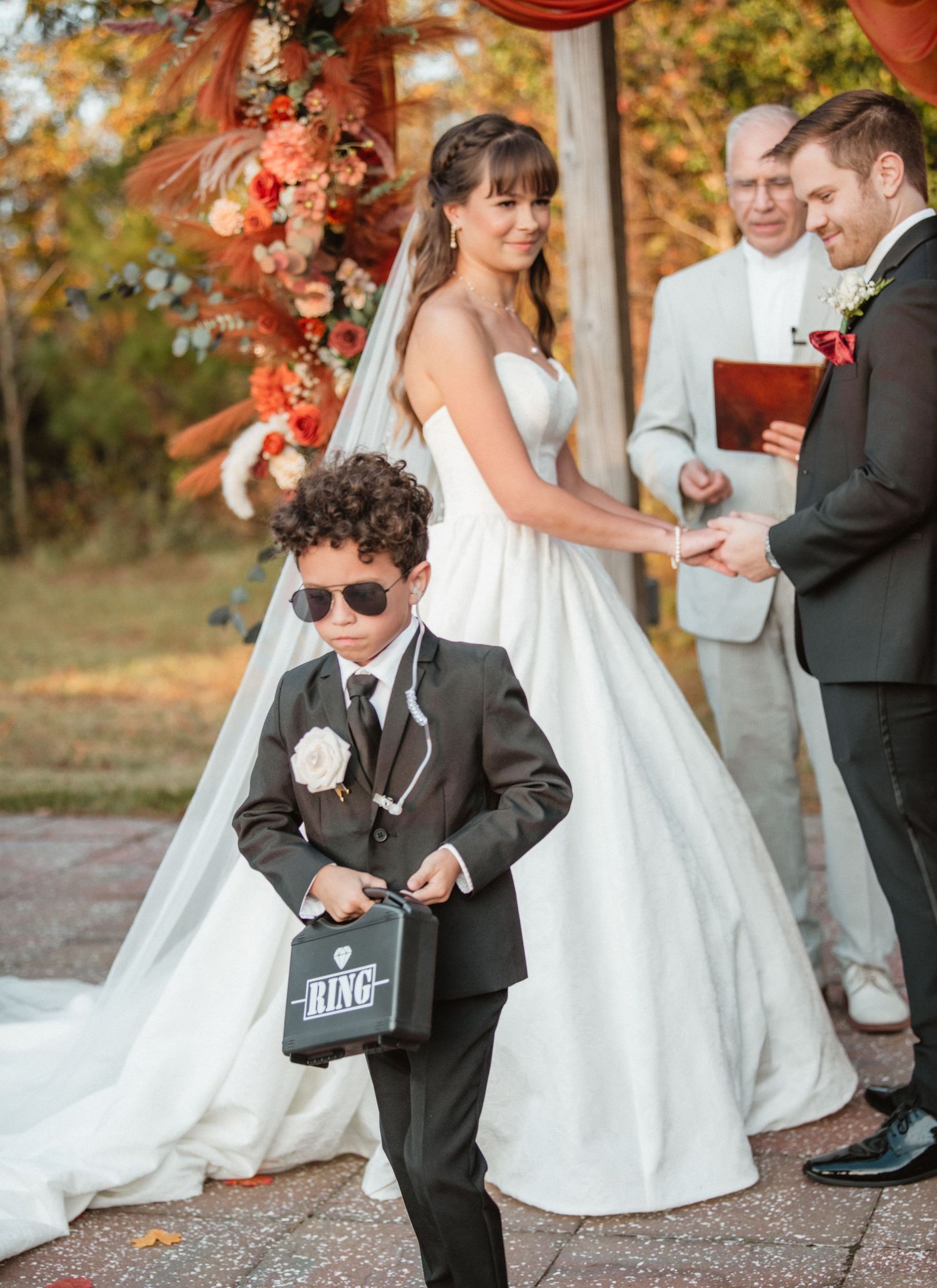 A young ring bearer in a black suit carries a sign during an outdoor wedding ceremony.